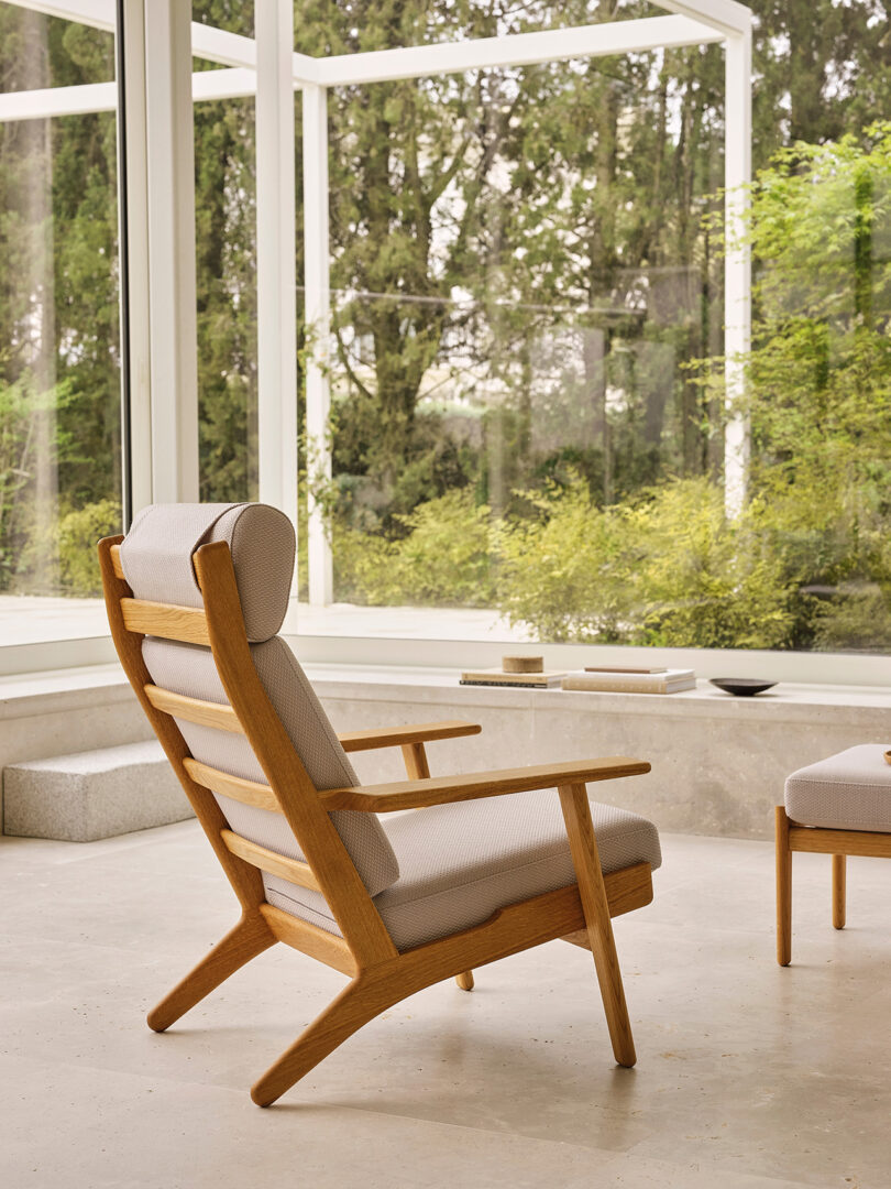 A wooden lounge chair with light-colored cushions sits on a stone floor in a glass-walled room overlooking green trees and bushes.