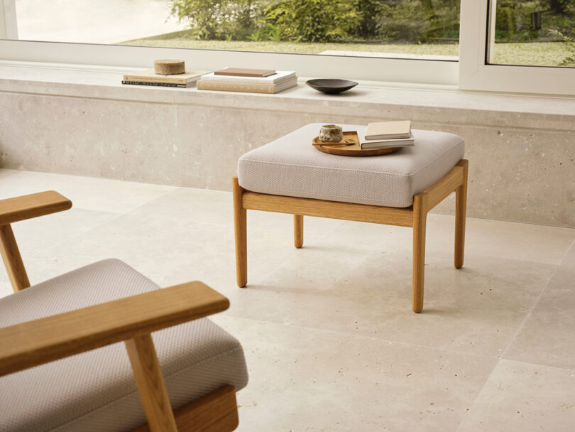 A cushioned wooden stool with a cup, saucer, and open notebook sits by a window, next to a matching armchair in a minimalist, light-filled room.