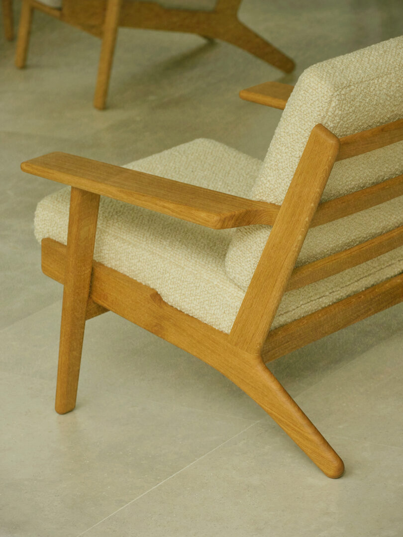 A wooden armchair with light textured upholstery is positioned on a light-colored tiled floor. Another similar chair is partially visible in the background.