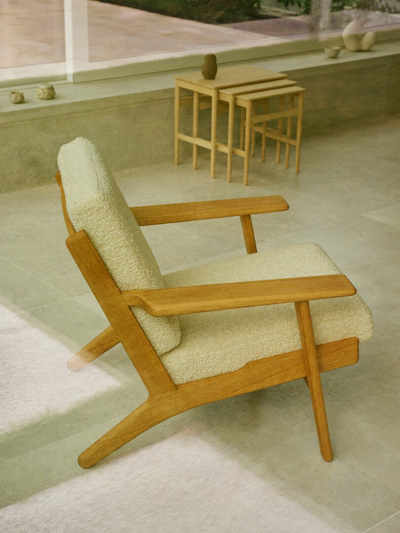 A wooden armchair with light beige upholstery is positioned in front of a window; nesting tables are visible in the background.