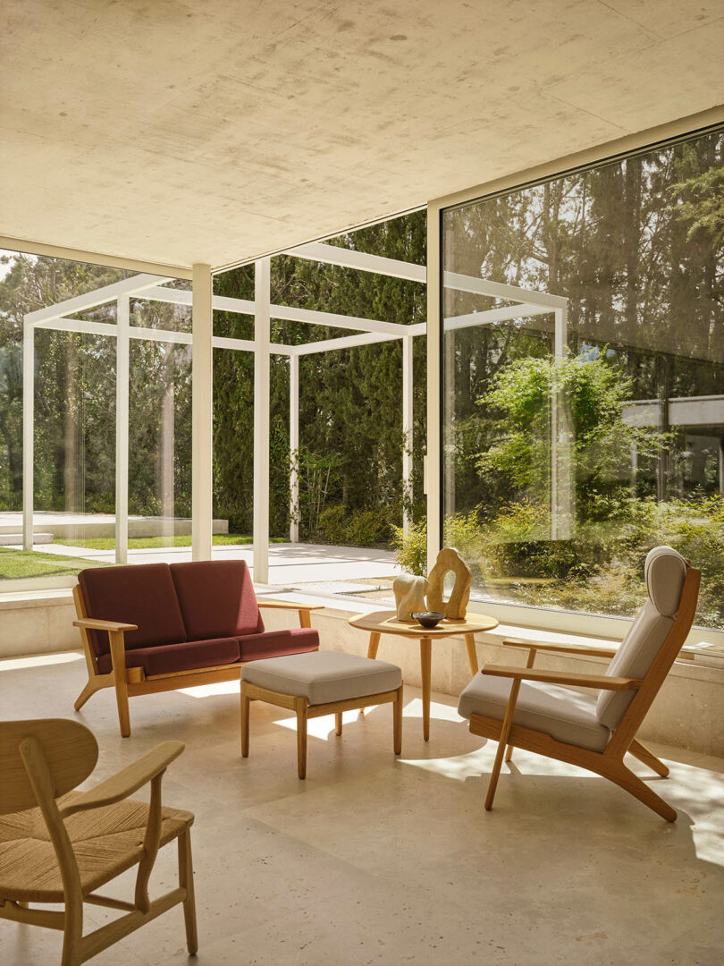 A modern sunroom with wooden furniture, large floor-to-ceiling windows, and views of greenery outside.