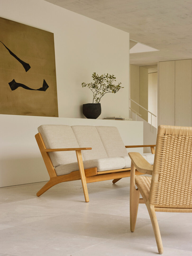 A modern minimalist living room with a wooden-framed beige sofa, a woven chair, a potted plant, and abstract wall art on a light-colored backdrop.