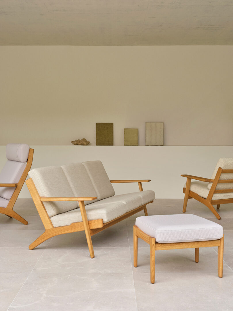 A modern living room with light wooden furniture, including a sofa, armchairs, and an ottoman, upholstered in beige fabric, set against a minimalist white wall with decorative objects.