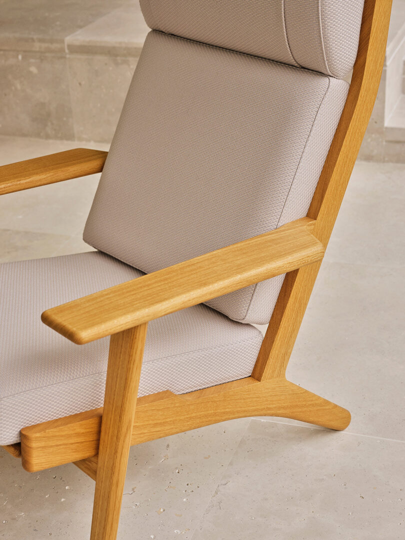 A close-up of a modern wooden armchair with light beige upholstered cushions, positioned on a stone tile floor.