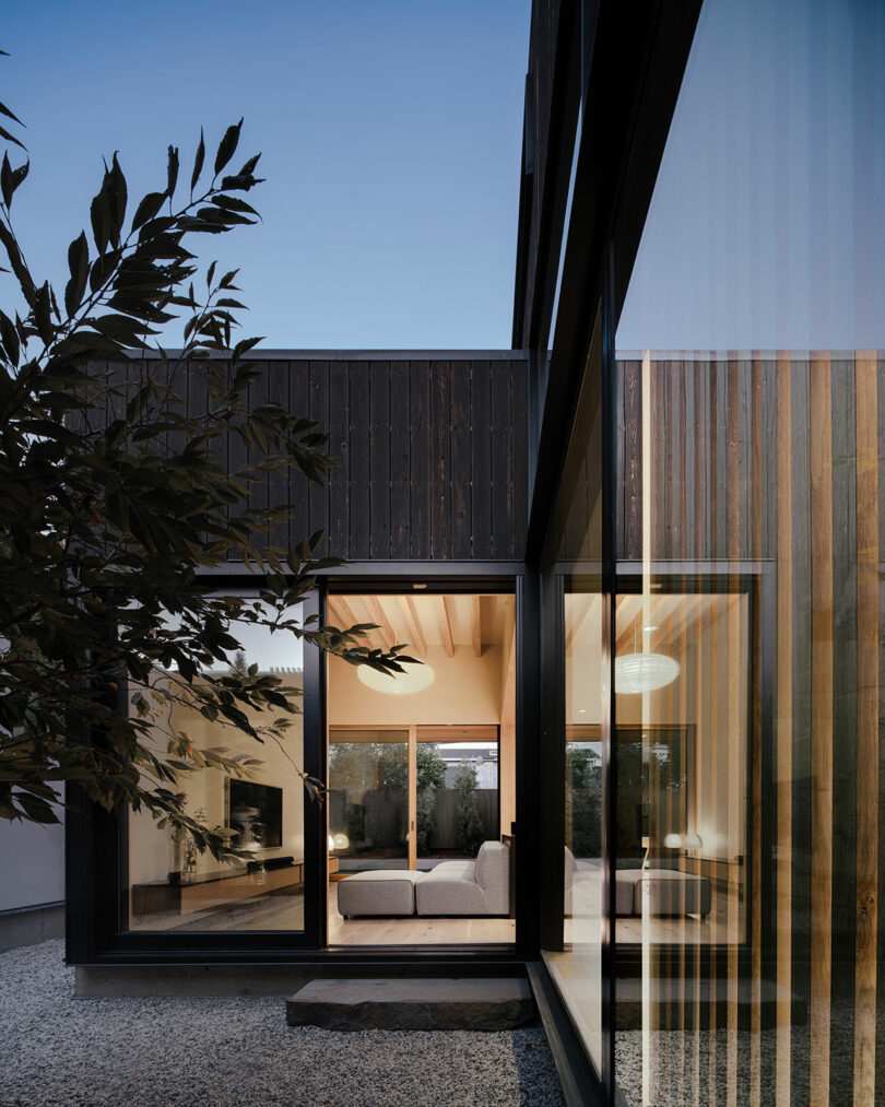 Modern house exterior with large glass windows, wooden and dark panel siding, and a view into a warmly lit living room. Gravel ground and a tree branch are visible in the foreground.