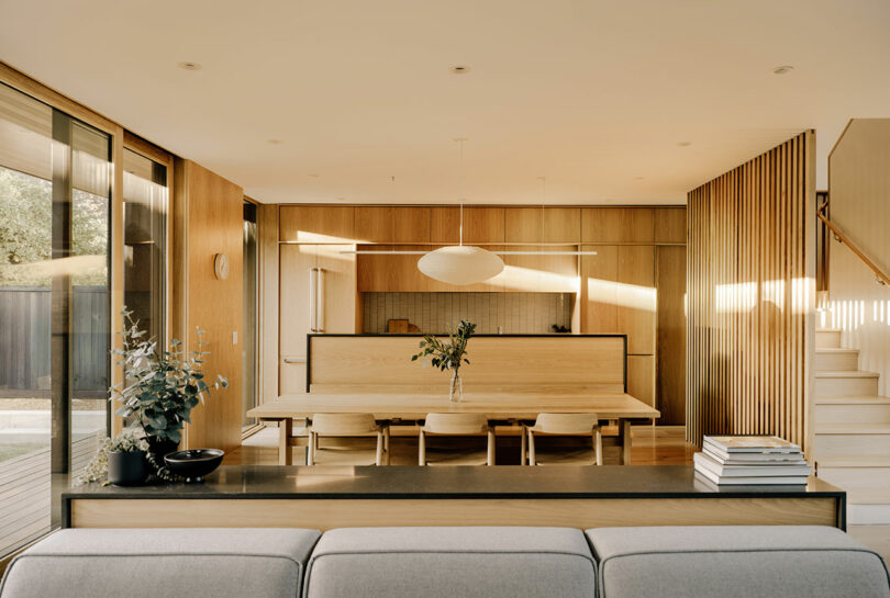 Modern open-plan living and dining area with wooden finishes, a light wood dining table, pendant light, and large windows letting in natural light.