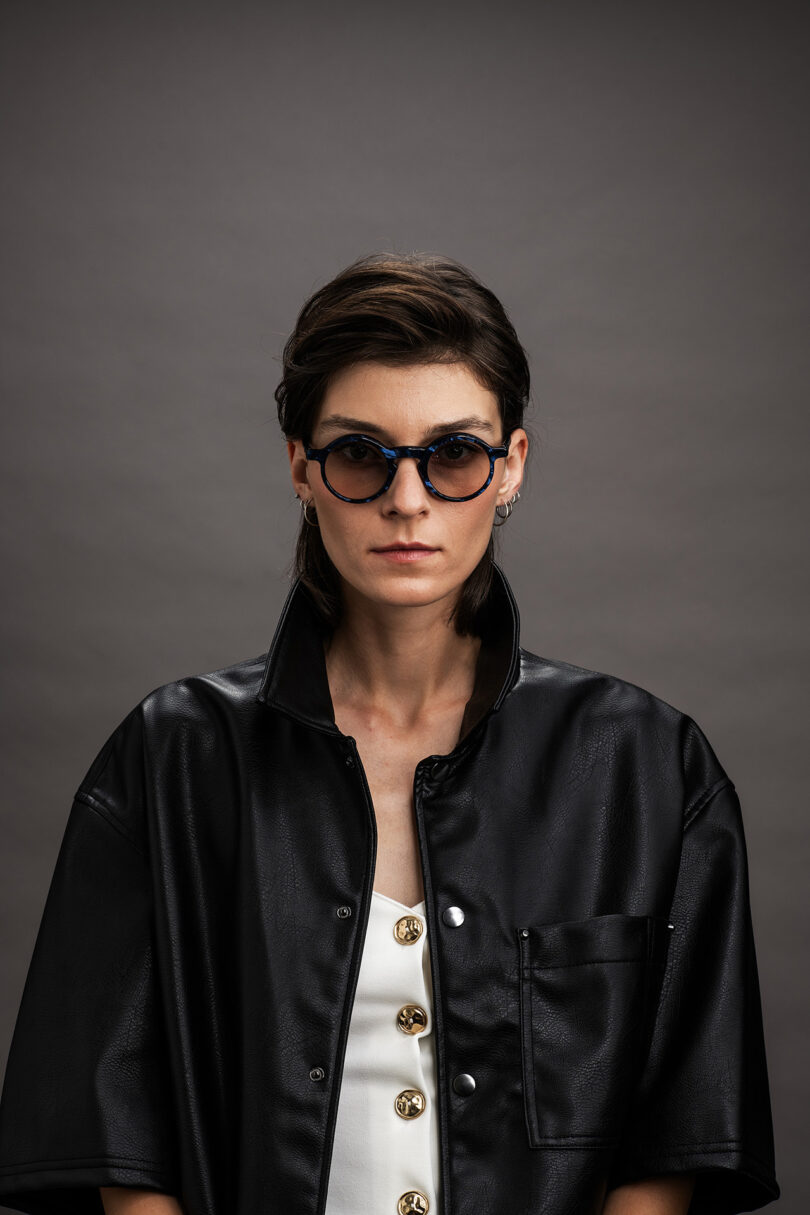 A woman with short dark hair wears round sunglasses, a black faux leather jacket with an open collar and a white top with gold buttons, standing against a gray background.