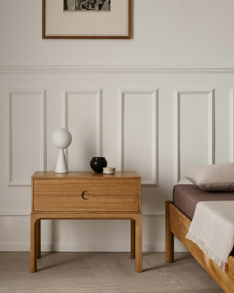 Wooden nighttliness with a round lamp, small black vase and two complex dishes - reminiscent of the design of Entra Kai Kristiansen next to the neutral bedding on the white coated wall.