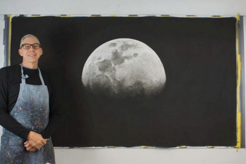 Una persona que usa anteojos y un delantal se encuentra junto a una gran pintura en blanco y negro detallada de la luna en una pared.