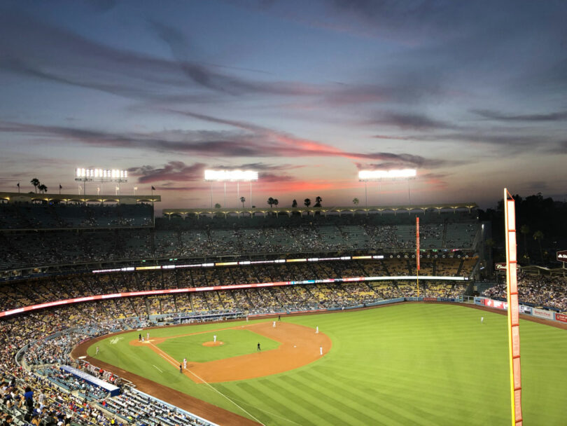 Un juego de béisbol en marcha en un estadio en la noche con el cielo mostrando colores de atardecer y luces del estadio que iluminan el campo.
