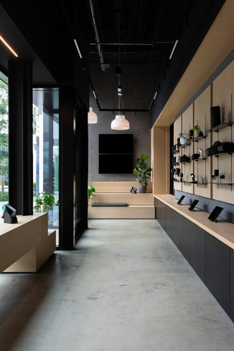 Interior de tiendas minoristas modernas con diseño minimalista, con dispositivos electrónicos que se muestran en estantes de pared, acentos de madera, piso de concreto y plantas en macetas por grandes ventanas.