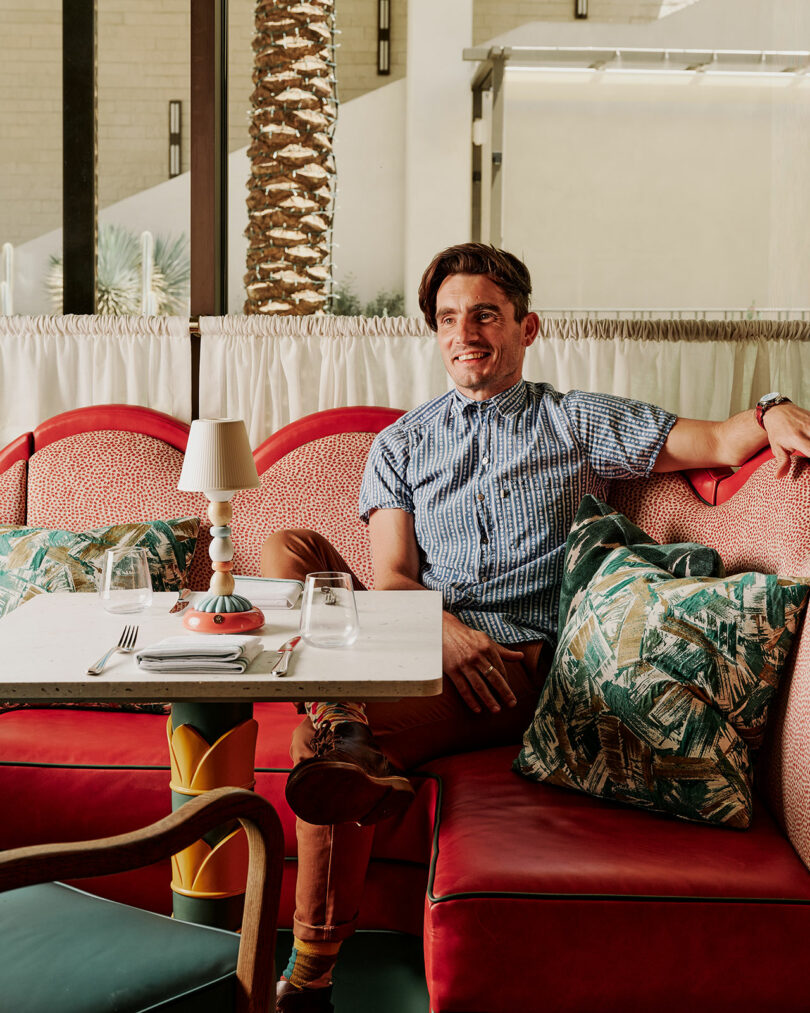 Tom Parker sits on a red patterned sofa in a restaurant, smiling with one arm on the backrest. A set table with glasses and cutlery is in front of him, and palm trees are visible outside.