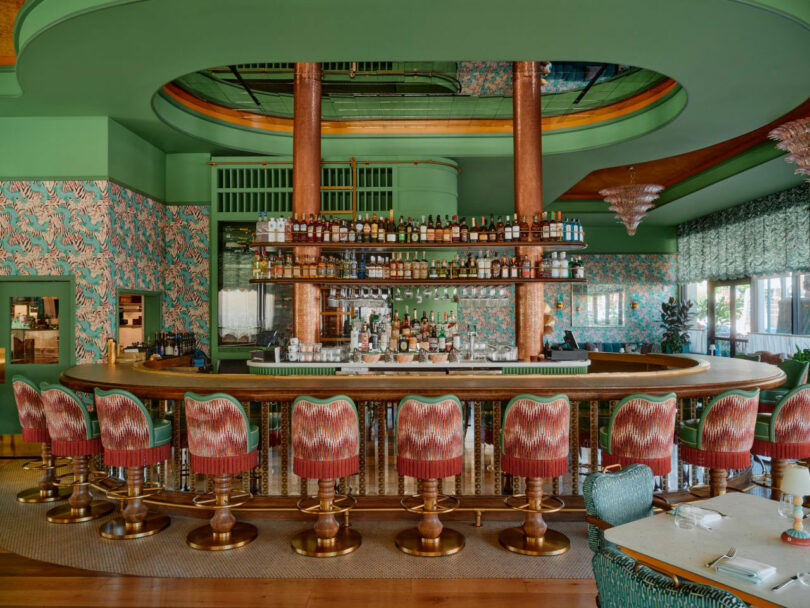 A curved bar with patterned upholstered stools, mirrored shelves holding bottles, green and pink patterned wallpaper, and green accents throughout the interior.