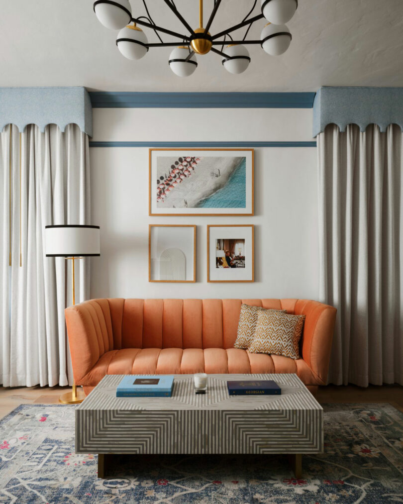 A modern living room with an orange sofa, patterned pillow, striped coffee table, floor lamp, framed wall art, chandelier, and long, light curtains.