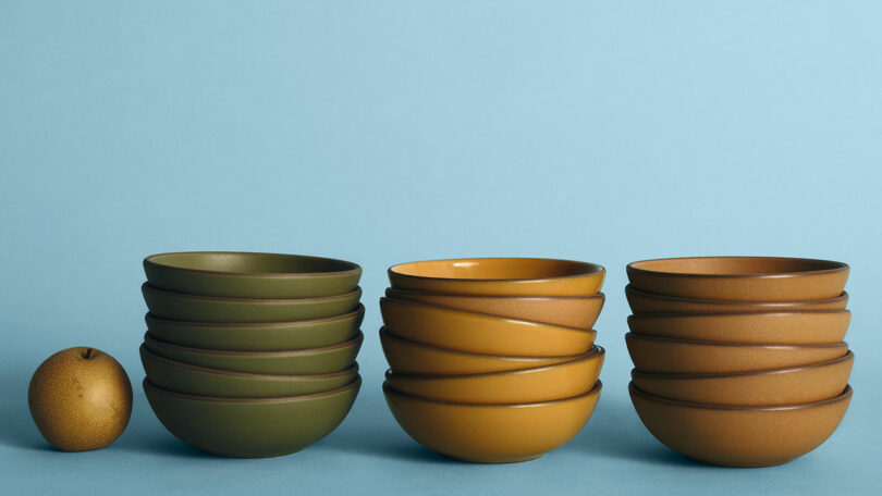 Three stacks of ceramic bowls in green, yellow, and brown are arranged on a blue surface, with a single brown Asian pear beside them against a blue background.