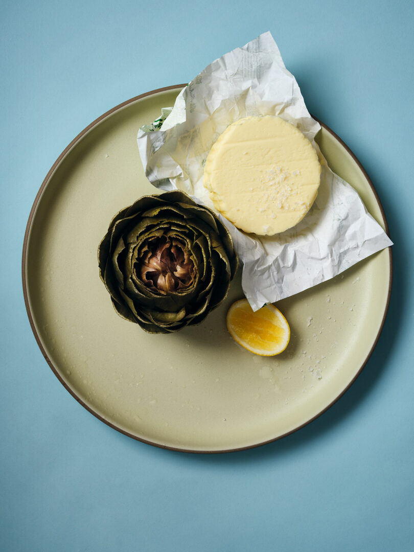 A plate with a whole artichoke, a pat of wrapped butter, and a lemon wedge on a light blue background.