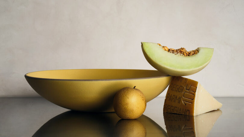 A yellow bowl, an Asian pear, a wedge of honeydew melon with seeds, and a block of cheese are arranged on a reflective surface against a neutral background.