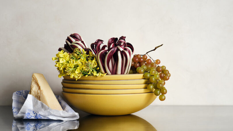 A yellow bowl holds radicchio, yellow flowers, and green grapes, with a wedge of cheese on blue-patterned paper beside it on a reflective surface.