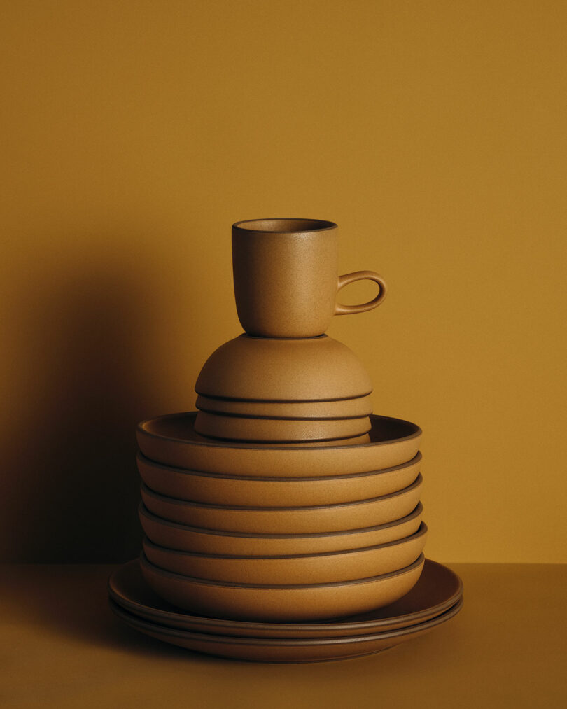 Stack of matte, earth-toned ceramic tableware including plates, bowls, and a cup, arranged neatly against a matching ochre background.