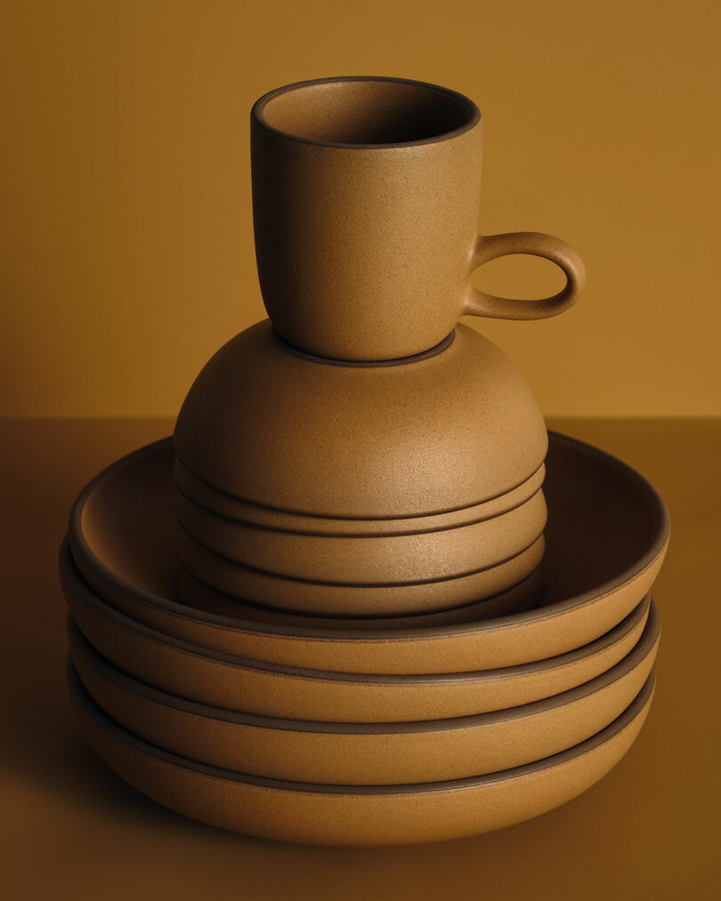 Stacked brown ceramic dishes and bowls with a matching cup on top, set against a solid brown background.