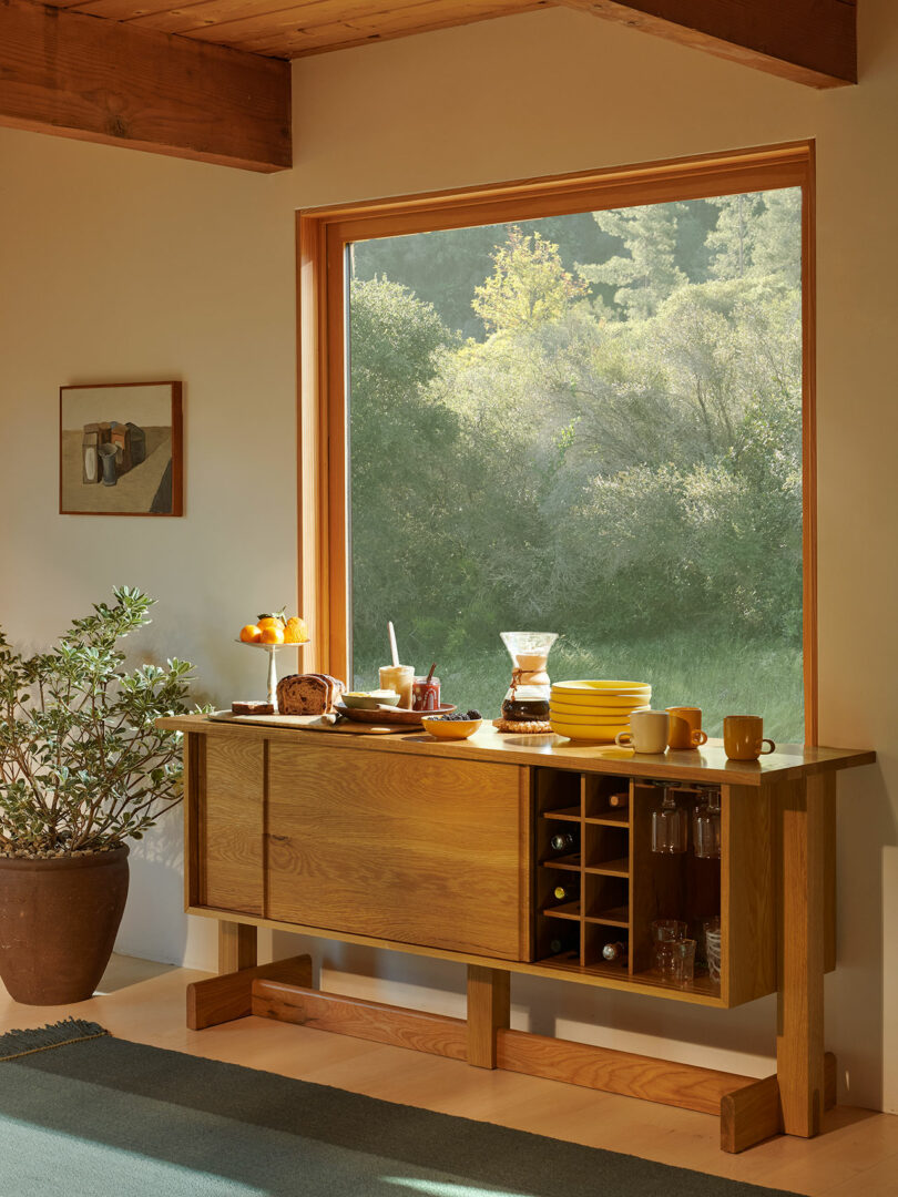 A wooden sideboard with breakfast items, dishes, a plant, and a coffee maker sits by a large window overlooking green trees in natural sunlight.