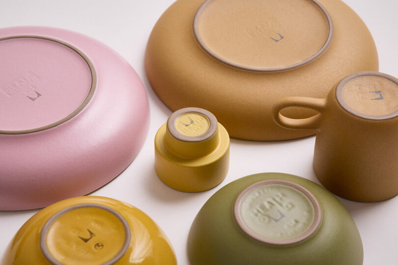 Gathered Heath Ceramics dishes, bowls, and a mug in pastel colors are arranged upside down, revealing their bases and maker's marks.
