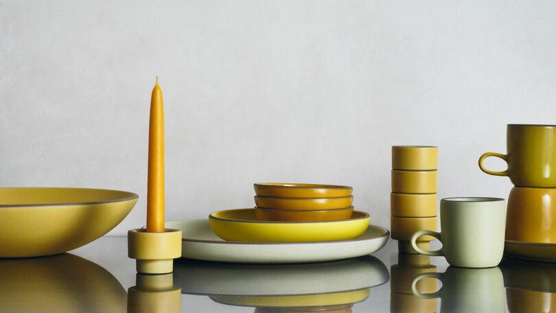 A collection of yellow and green ceramic tableware, including bowls, plates, mugs, and a candle, arranged on a reflective surface against a light background.