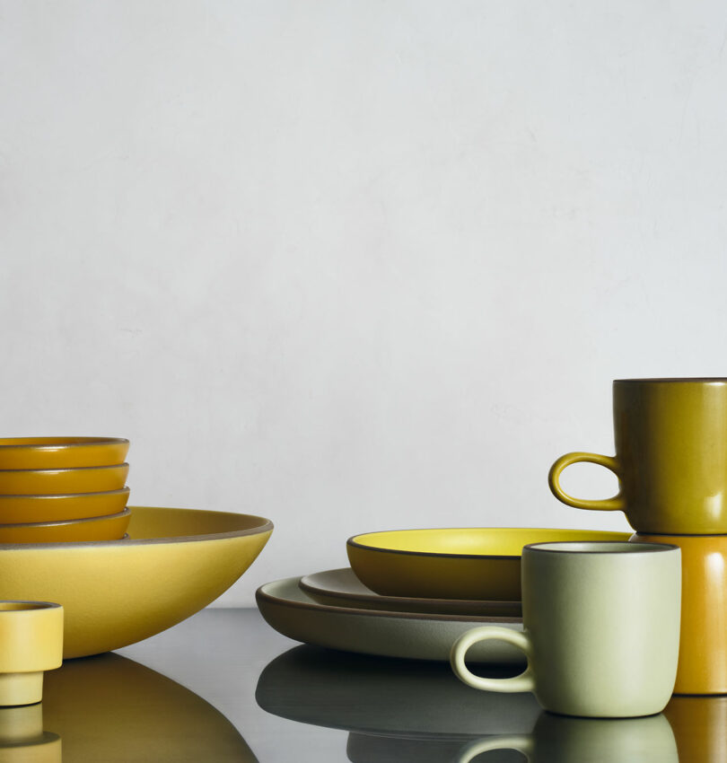 A collection of ceramic bowls, plates, and mugs in yellow, green, and beige tones arranged on a reflective surface against a plain light background.