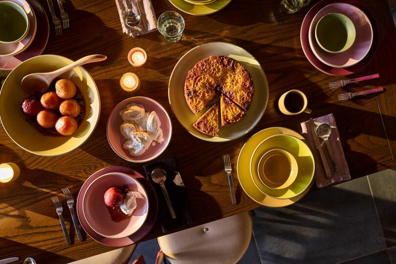 Overhead view of a set dining table with plates, cups, utensils, candles, a cake with a slice cut, baked apples, and a bowl of ice cream.