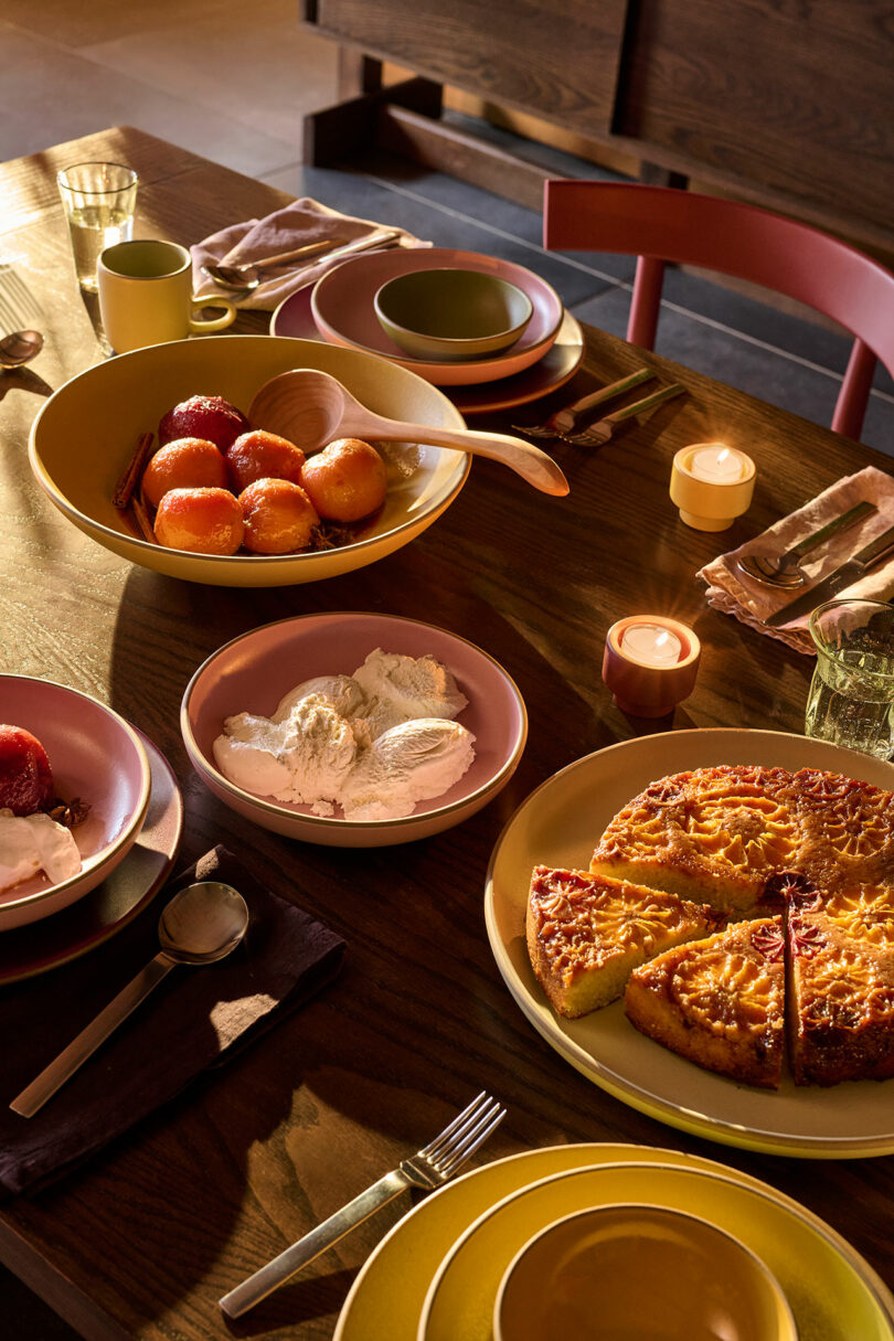 A wooden table set for dessert with plates, cutlery, candles, a bowl of apples, a bowl of ice cream, and a sliced cake.