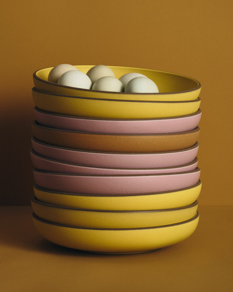 A stack of yellow and pink bowls holds several eggs against a solid mustard-colored background.