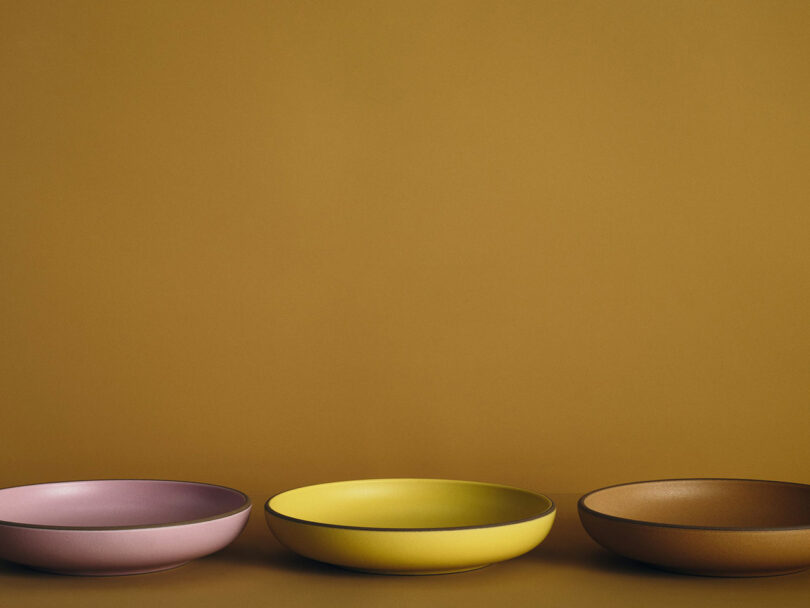 Three shallow bowls in pink, yellow, and brown are arranged side by side against a plain mustard-colored background.