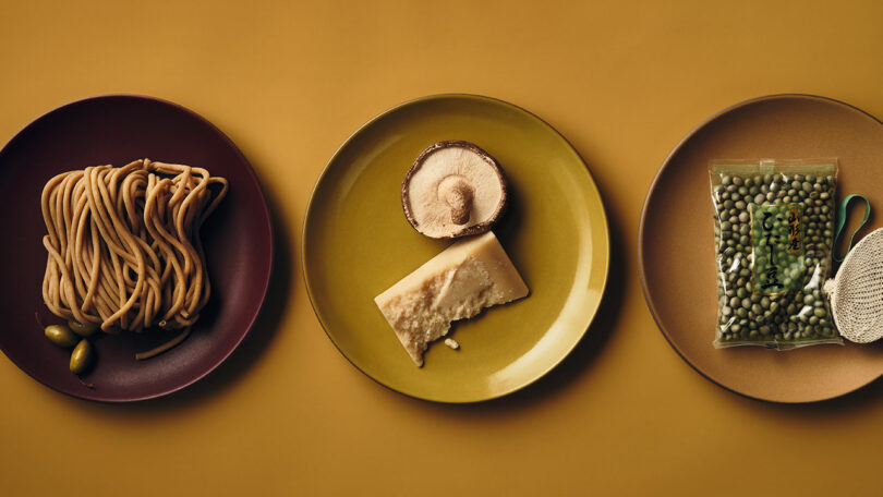 Three plates on a yellow surface: one with noodles and olives, one with a mushroom and parmesan cheese, and one with a vacuum-sealed bag of green lentils and a loofah.