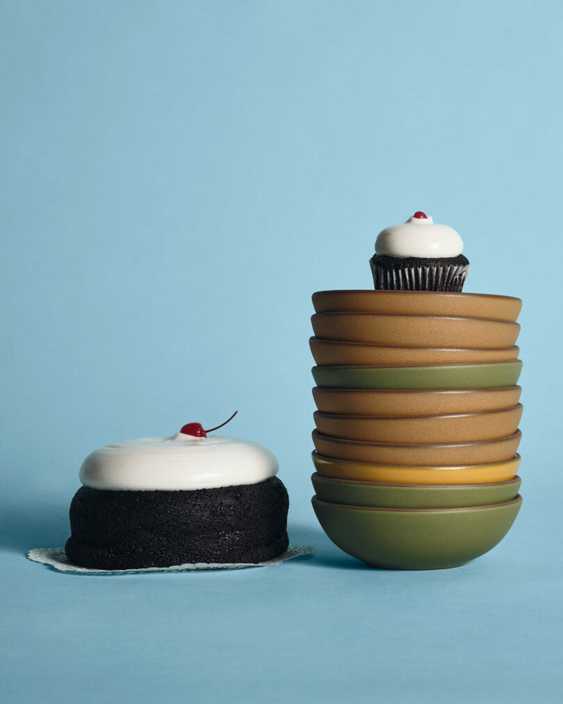 A chocolate cake with white frosting and a cherry is next to stacked bowls topped with a cupcake decorated the same way, all set against a blue background.