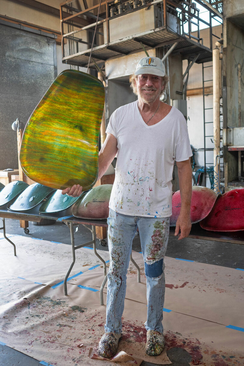 A man in paint-splattered clothes and a white hard hat stands in a workshop, holding a large, green and yellow sculpted object. Several similar objects are on tables behind him.