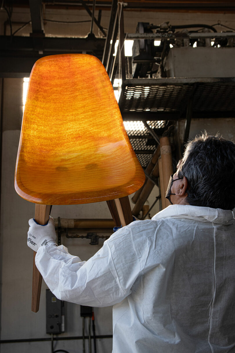 A person in protective clothing holds up an amber-colored translucent chair with wooden legs, examining it under industrial lighting in a workshop.