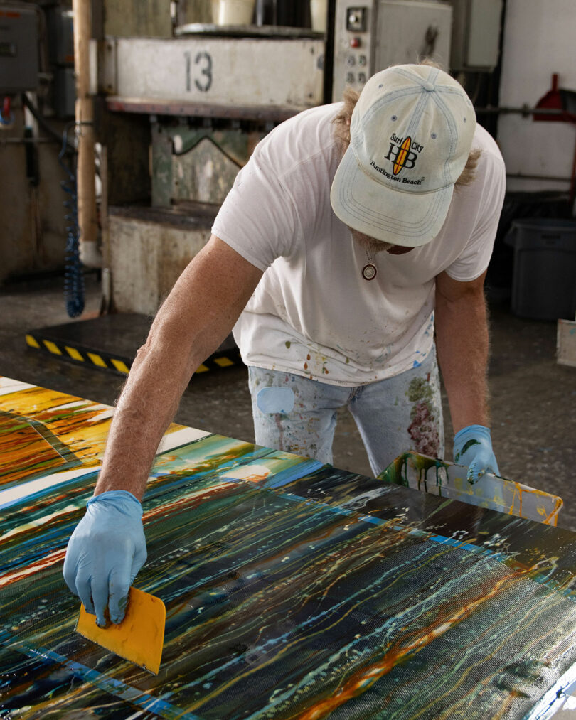 A person wearing gloves and a cap spreads paint across a large canvas with a yellow tool in an industrial studio setting.