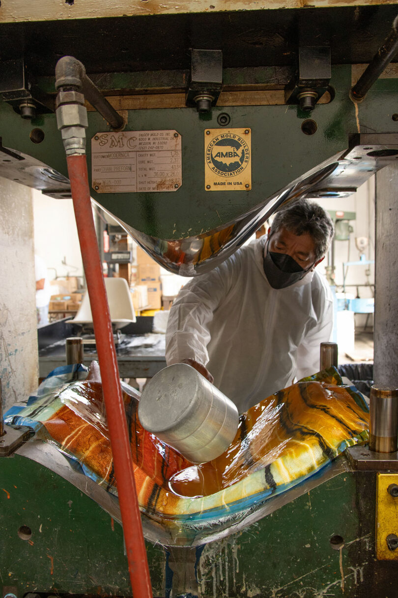 A worker wearing protective gear pours resin onto colorful fiberglass sheets inside an industrial press at a manufacturing facility.