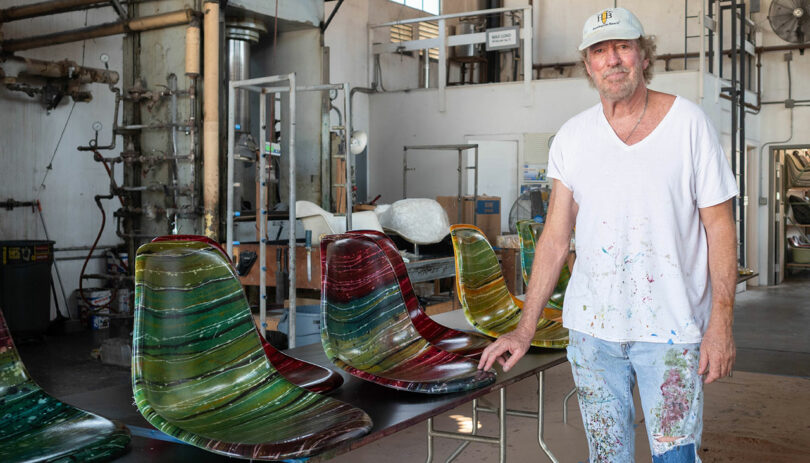 A man in paint-splattered clothes stands next to a table displaying colorful, striped chair seats in an industrial workshop.
