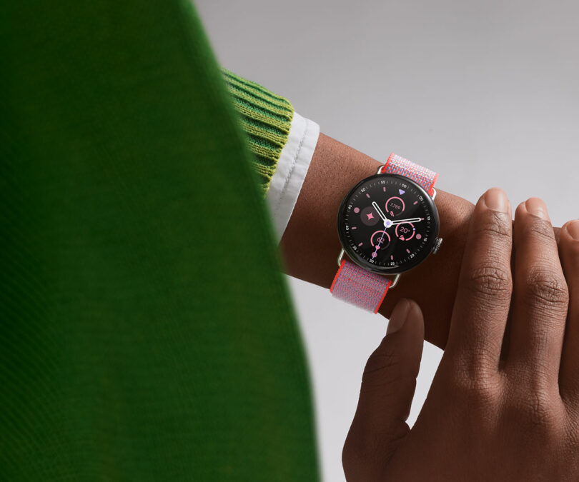 A person wearing a smart watch with a round black face and a pink-and-white woven strap, touching the watch with their other hand. The person is dressed in a green sweater.