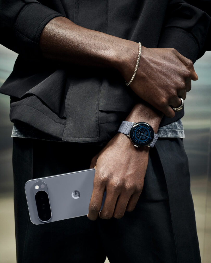 A person wearing a dark outfit holds a silver Google Pixel phone in one hand and wears a smartwatch with a round blue face on the opposite wrist.