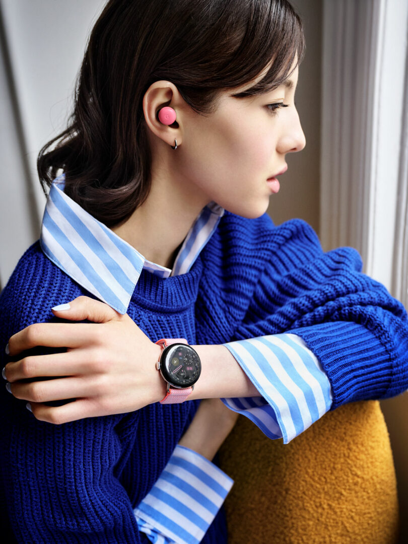 Woman in blue sweater wearing a smartwatch and pink wireless earbud, sitting by a window with arms crossed, looking outside.