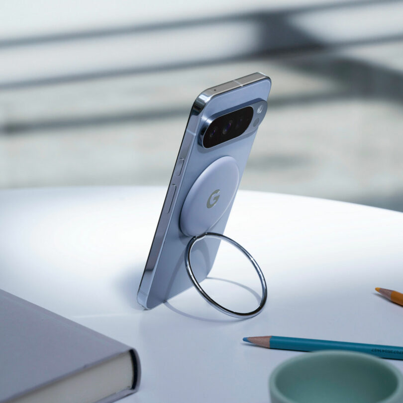 A smartphone with a circular metal ring stand attached to the back is propped upright on a white table next to a notebook, pencil, and a green cup.