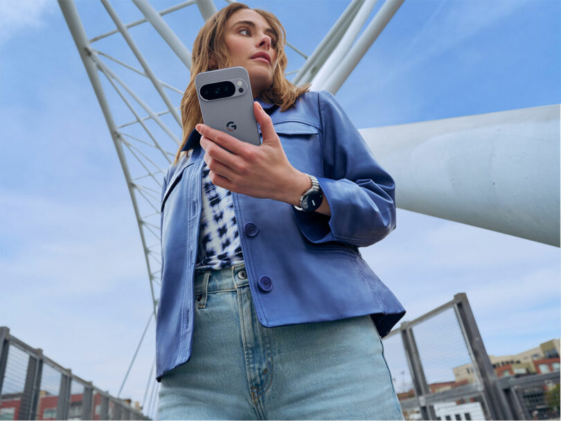 A woman in a blue jacket and jeans holds a Google Pixel smartphone while standing outdoors under a large metal structure.