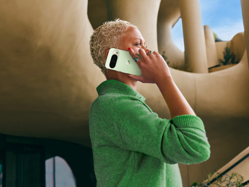 A person wearing a green sweater talks on a mint green smartphone while standing outside a modern, curved architectural building.