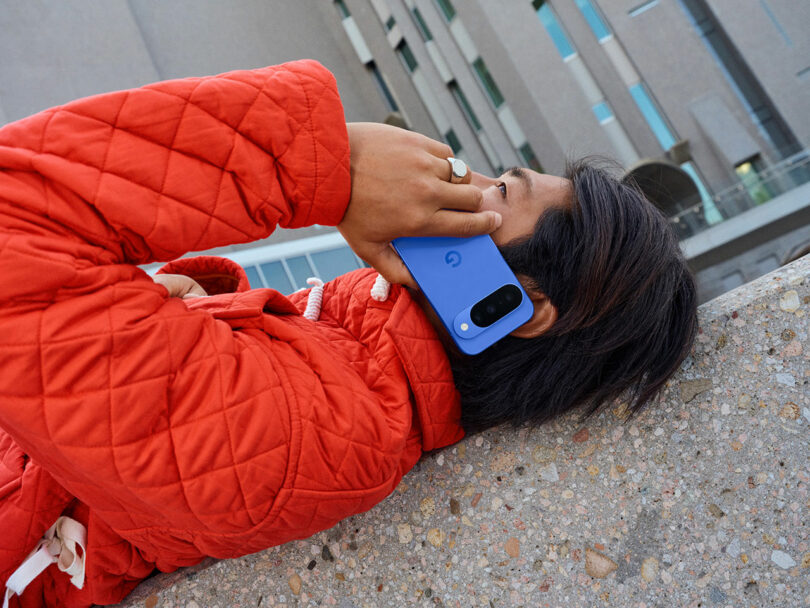 A person in a red quilted jacket is lying on a concrete surface, holding a blue smartphone to their ear.