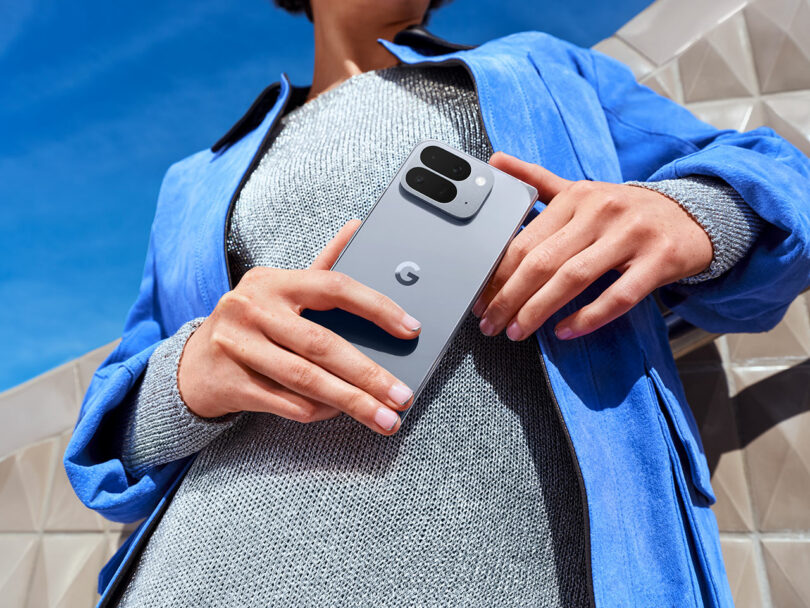 A person wearing a blue jacket and gray top holds a Google Pixel smartphone outdoors, with an architectural geometric structure in the background.