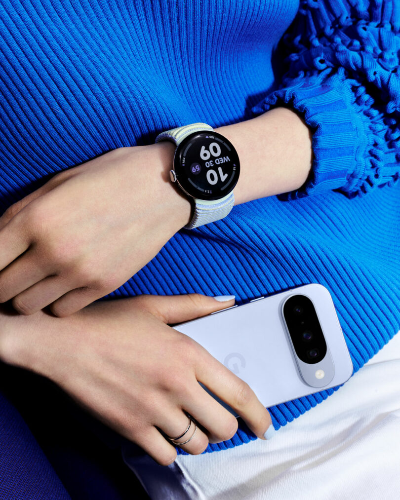 A person wearing a blue sweater holds a white smartphone and wears a smartwatch showing the time as 10:50 on their left wrist.