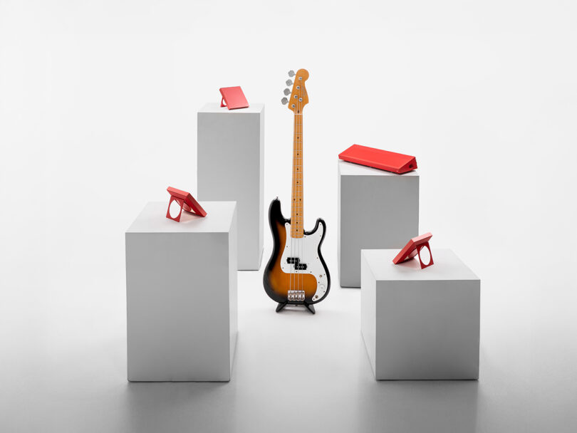 A black-and-white electric bass guitar stands upright in the center, surrounded by four white pedestals with red tablets on top, against a plain white background.