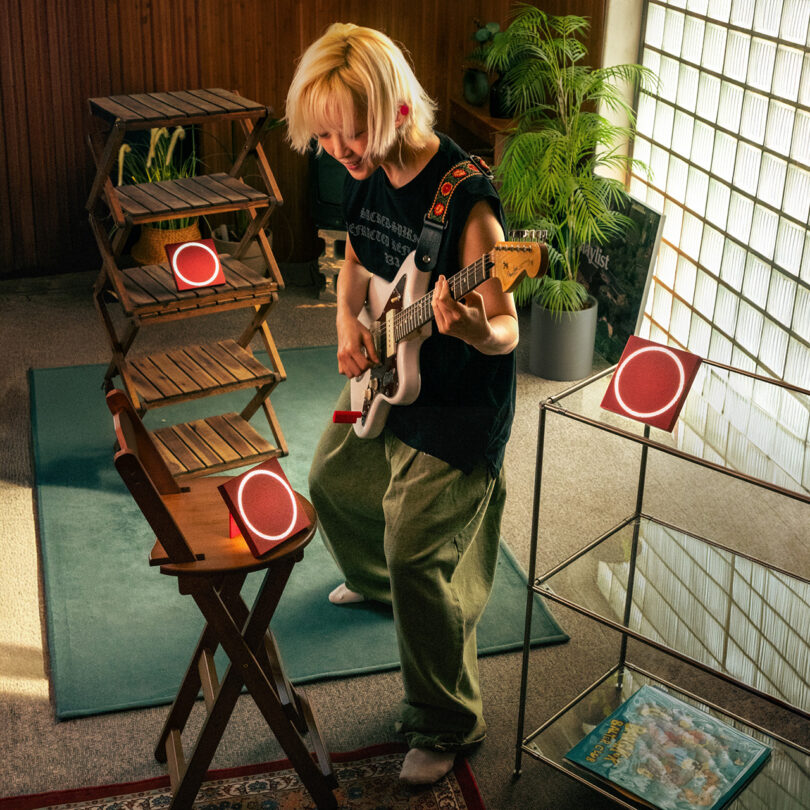 A person with a guitar stands in a room with wooden shelves, small red speakers, a plant, and a magazine on a metal rack.